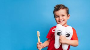 Little boy holding a crocheted tooth plush and a wooden toothbrush in front of a blue background